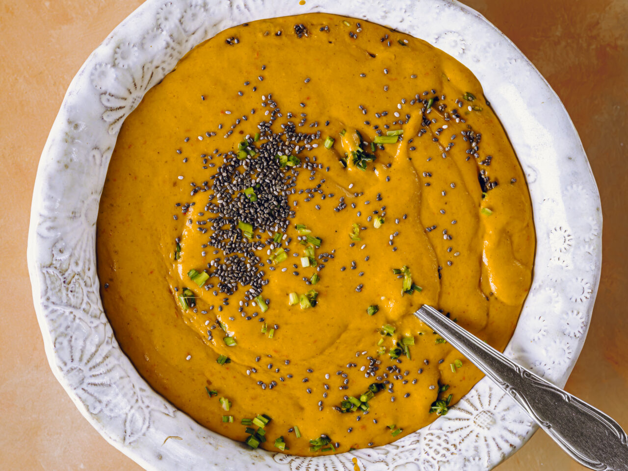 Bowl of creamy orange vegetable soup topped with chia seeds and chopped chives, with a spoon resting in the bowl.