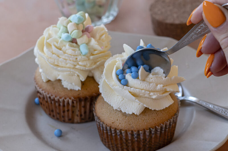 Finished muffins topped with whipped cream and pastel sprinkles on a plate, a spoon diving in.