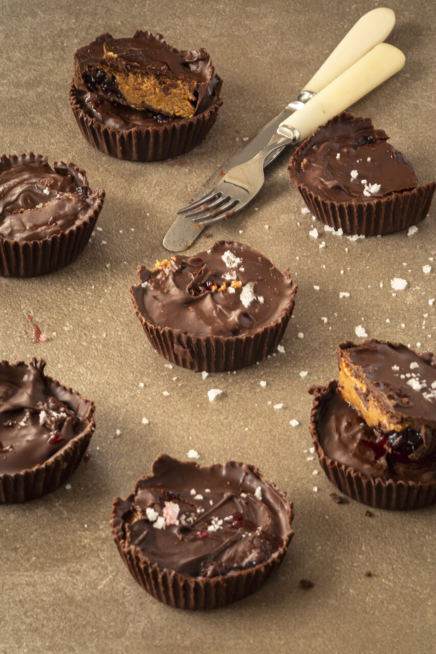 Homemade chocolate peanut butter cups scattered on a surface, some cut open to reveal peanut butter and jam filling, sprinkled with flaky sea salt and served with a small fork and knife.