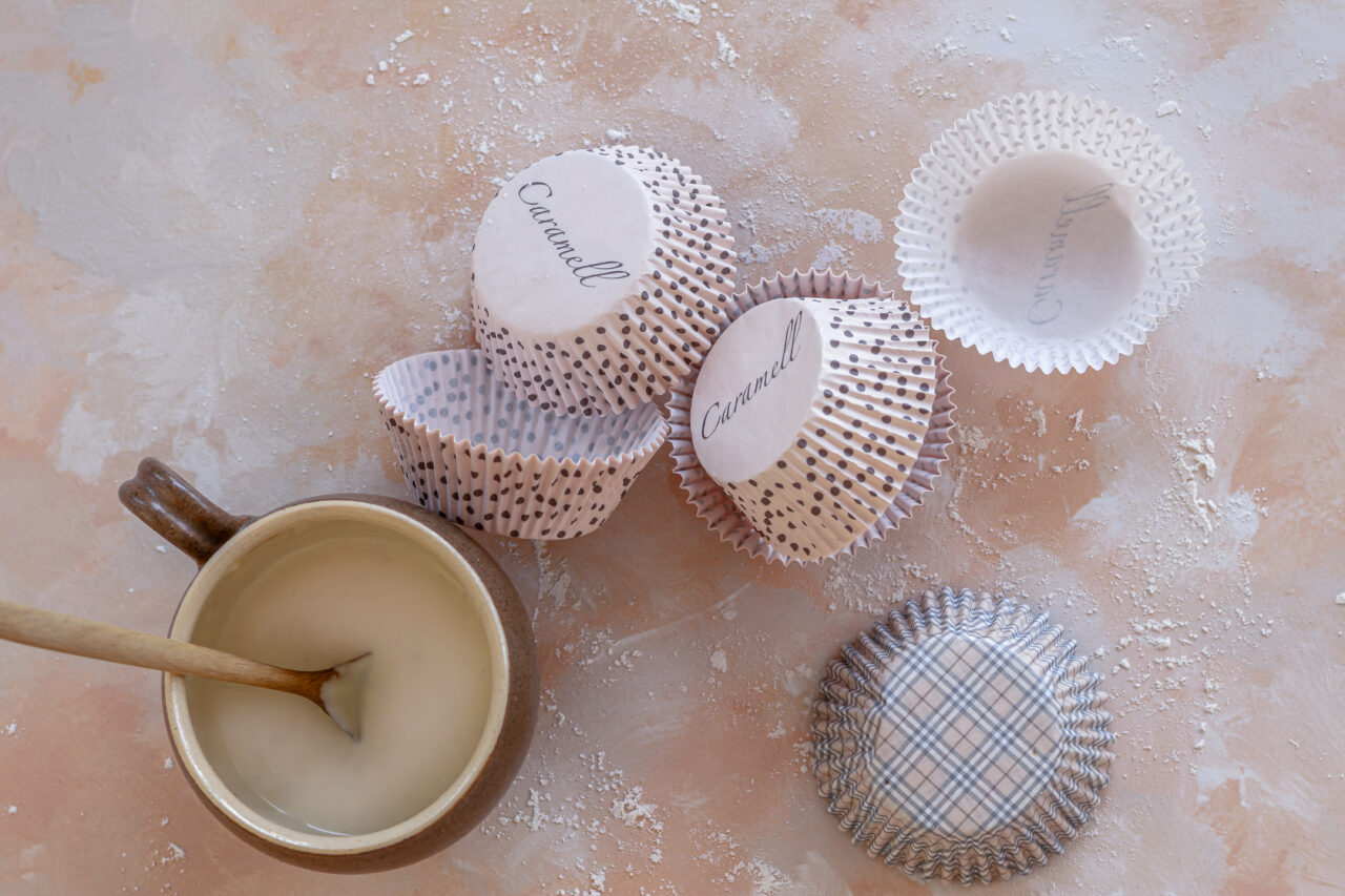 Paper muffin liners and a small cup of melted white chocolate on a lightly floured surface, baking setup.