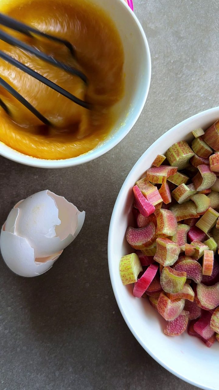 Whisking eggs and sugar in a bowl next to a bowl of chopped rhubarb