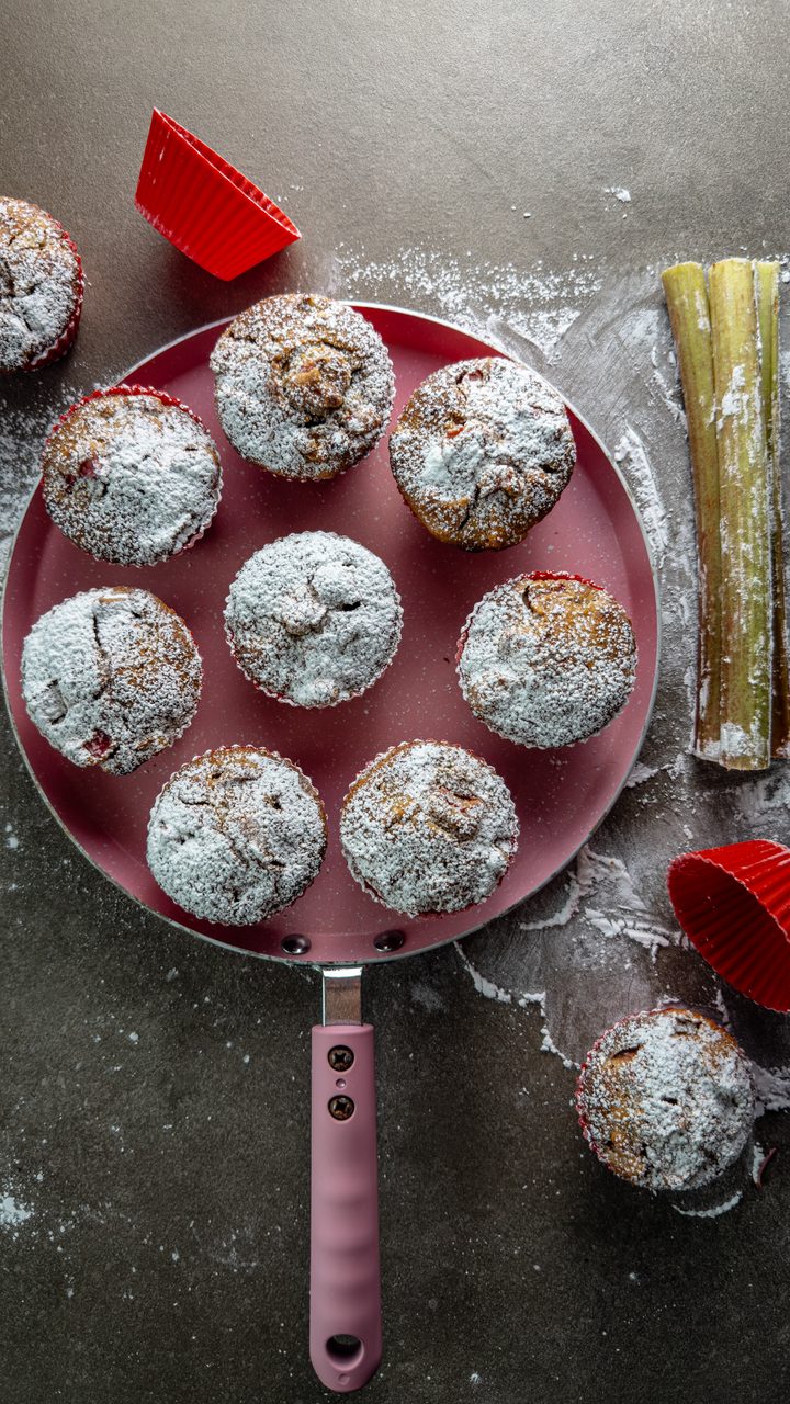 Eight rhubarb muffins dusted with icing sugar on a pink pan, with fresh rhubarb stalks and red silicone muffin cups beside it on a dark surface