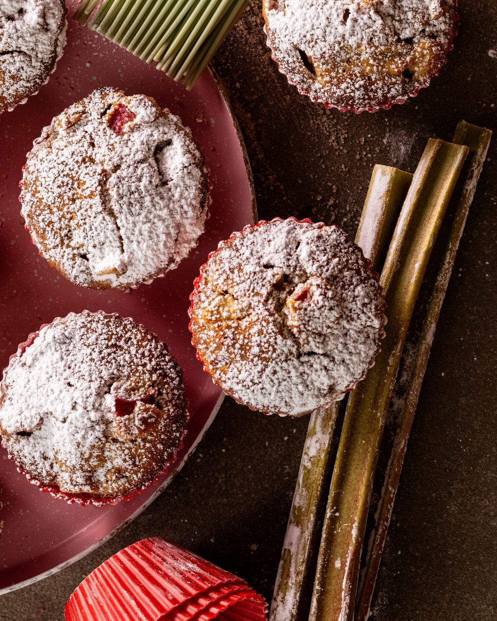 Sourdough rhubarb muffins dusted with icing sugar on a pink plate, with fresh rhubarb stalks and red silicone muffin cups beside them