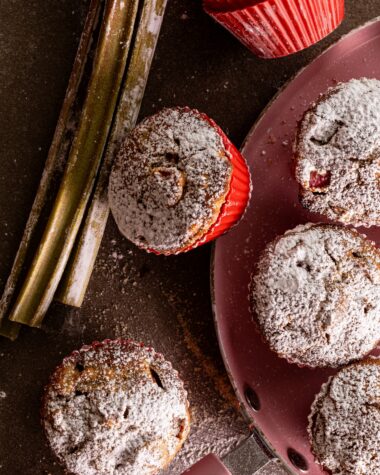 Sourdough rhubarb muffins dusted with icing sugar on a pink pan, next to fresh rhubarb stalks and a muffin in a red silicone cup