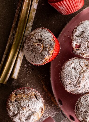 Sourdough rhubarb muffins dusted with icing sugar on a pink pan, next to fresh rhubarb stalks and a muffin in a red silicone cup