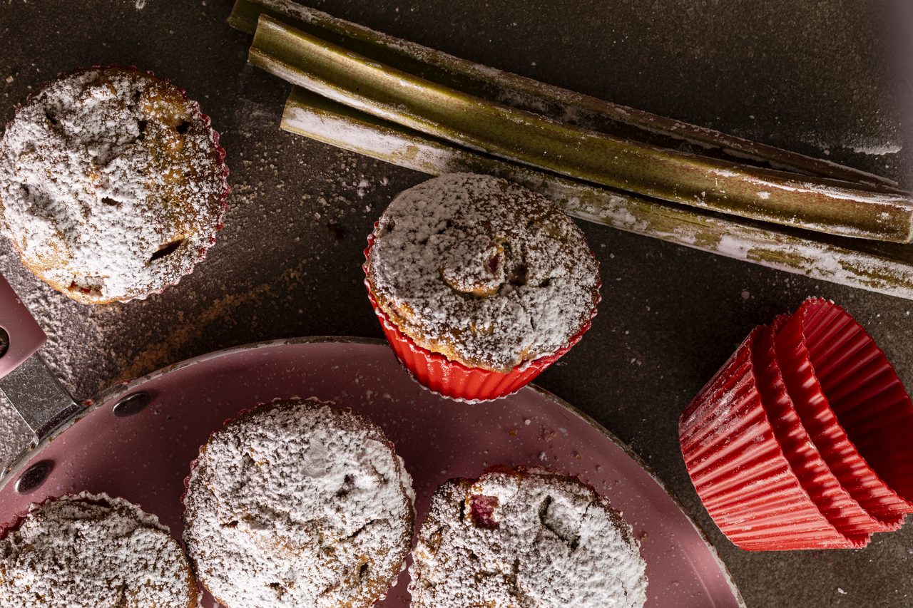 Overhead view of rhubarb muffins dusted with icing sugar on a pink pan, with one muffin in a red silicone cup, fresh rhubarb stalks and a stack of red muffin cups